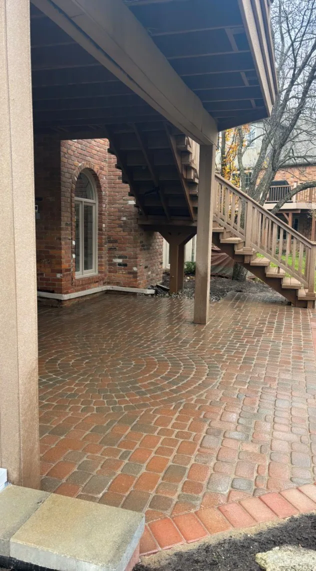 Covered paver patio beneath a raised deck, laid in a red cobblestone fan pattern against the brick wall of the house