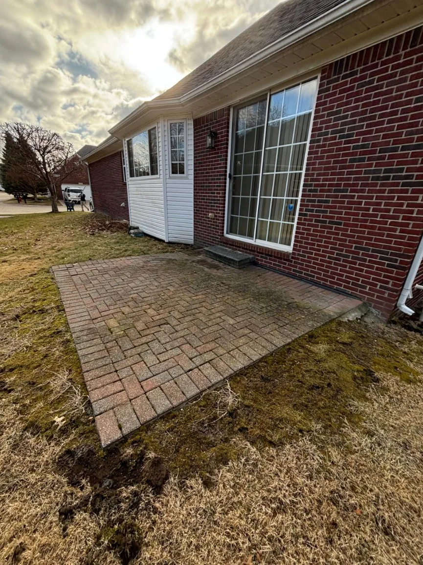 Herringbone brick paver patio in warm red and tan tones running alongside a red brick ranch home with a white bay window