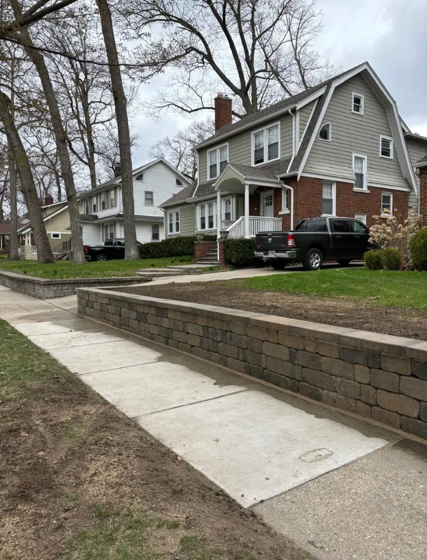 Gray segmental block retaining wall built along a concrete driveway and front walk, holding back the raised front lawn of a two-story home