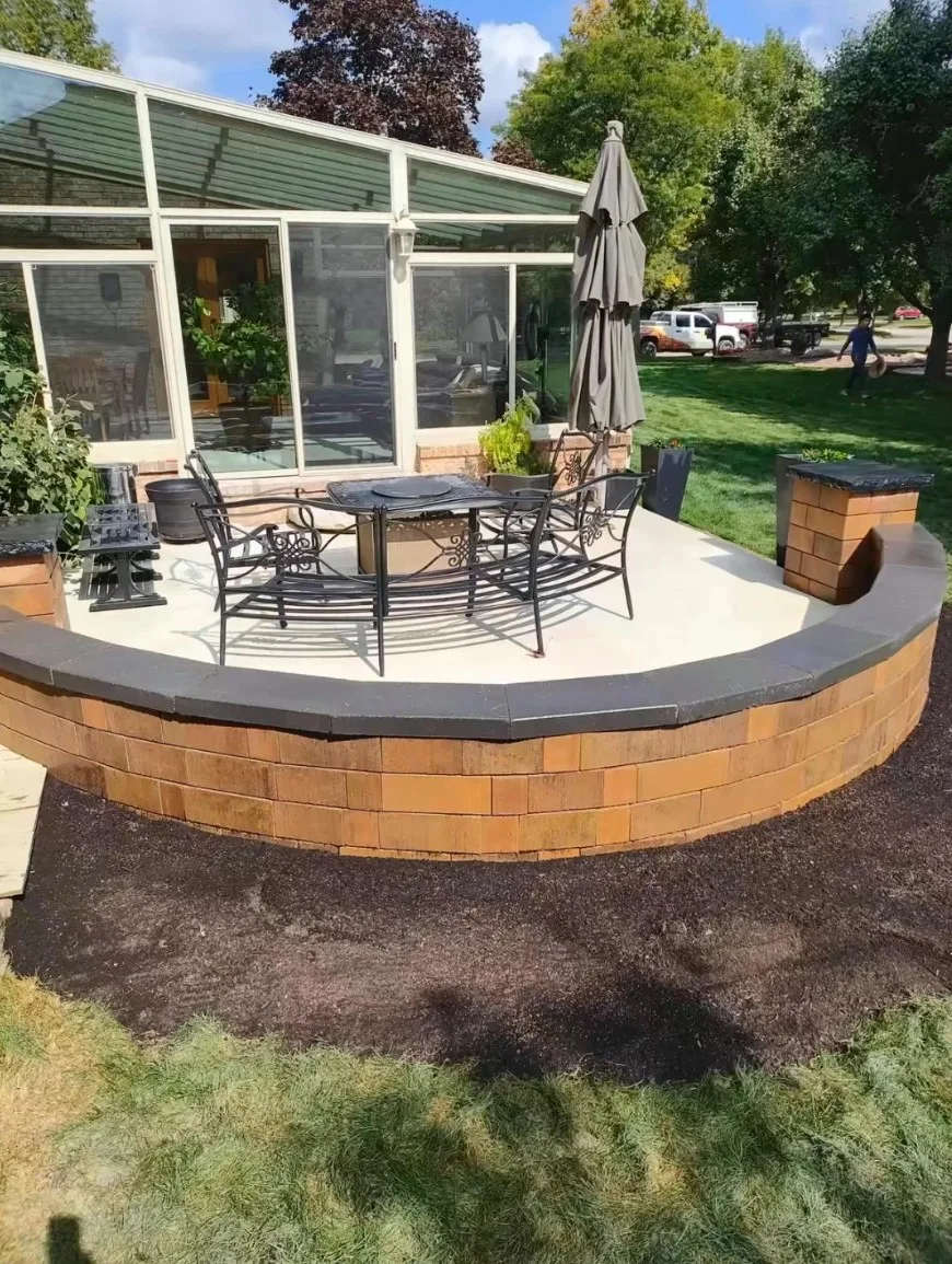 Curved raised patio with a tan block seating wall and gray coping caps, set with patio furniture beside a glass sunroom